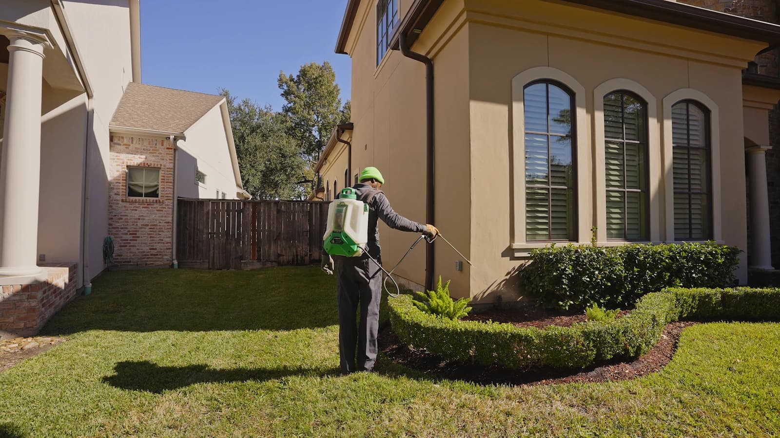 Technician with a backpack sprayer treating the foundation and mulched landscape bed of a two-story home