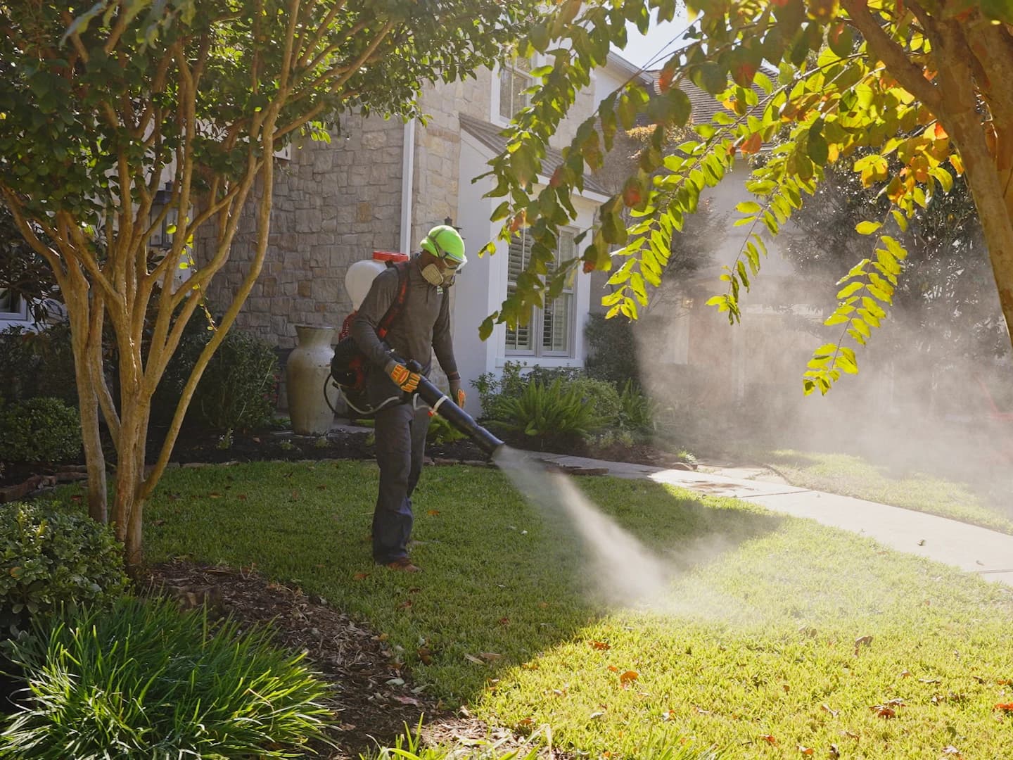 Technician in a respirator, gloves, and high-visibility hat applying a backpack mister treatment across a green residential lawn
